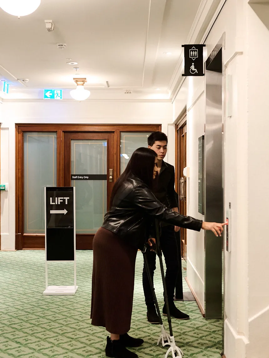A woman on crutches and a man stand outside a lift on green carpet in Old Parliament House.