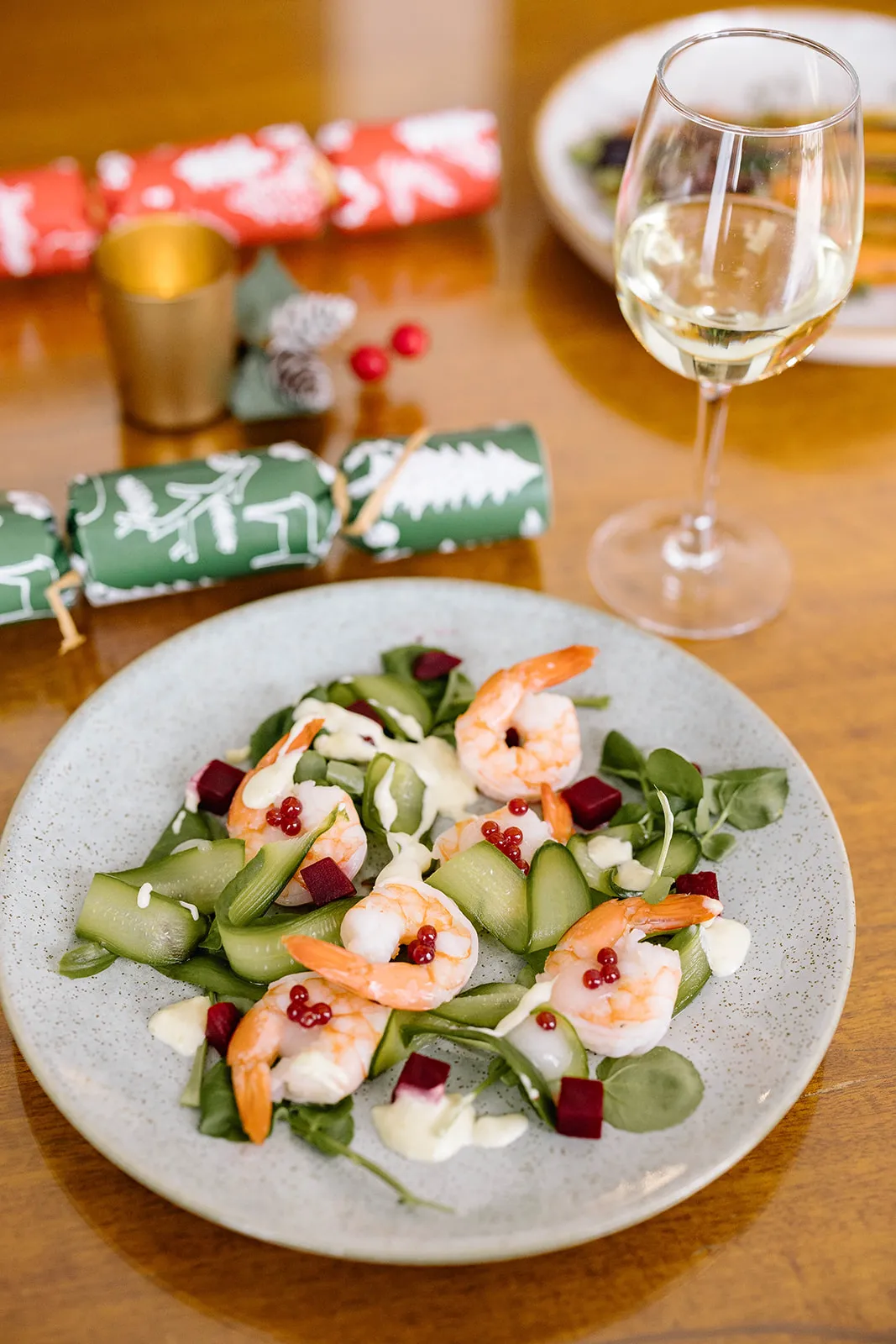 A grey plate with prawns and salad sits on top of a wood table. A green Christmas cracker and wine glass are beside the plate.