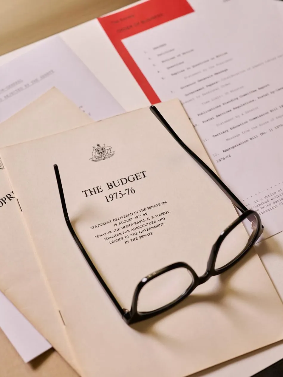 A stack of papers on a desk, a yellow booklet titled 'The Budget 1975-76' with the Commonwealth Coat of Arms is on top, along with a pair of black-framed reading glasses and a pen.