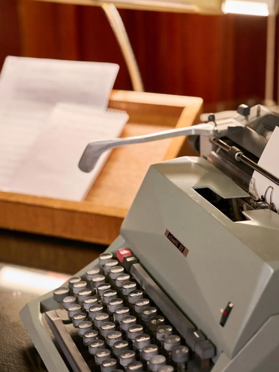 An old-fashioned grey typewriter and wooden document tray with paper in it sits on green leather-topped desk.