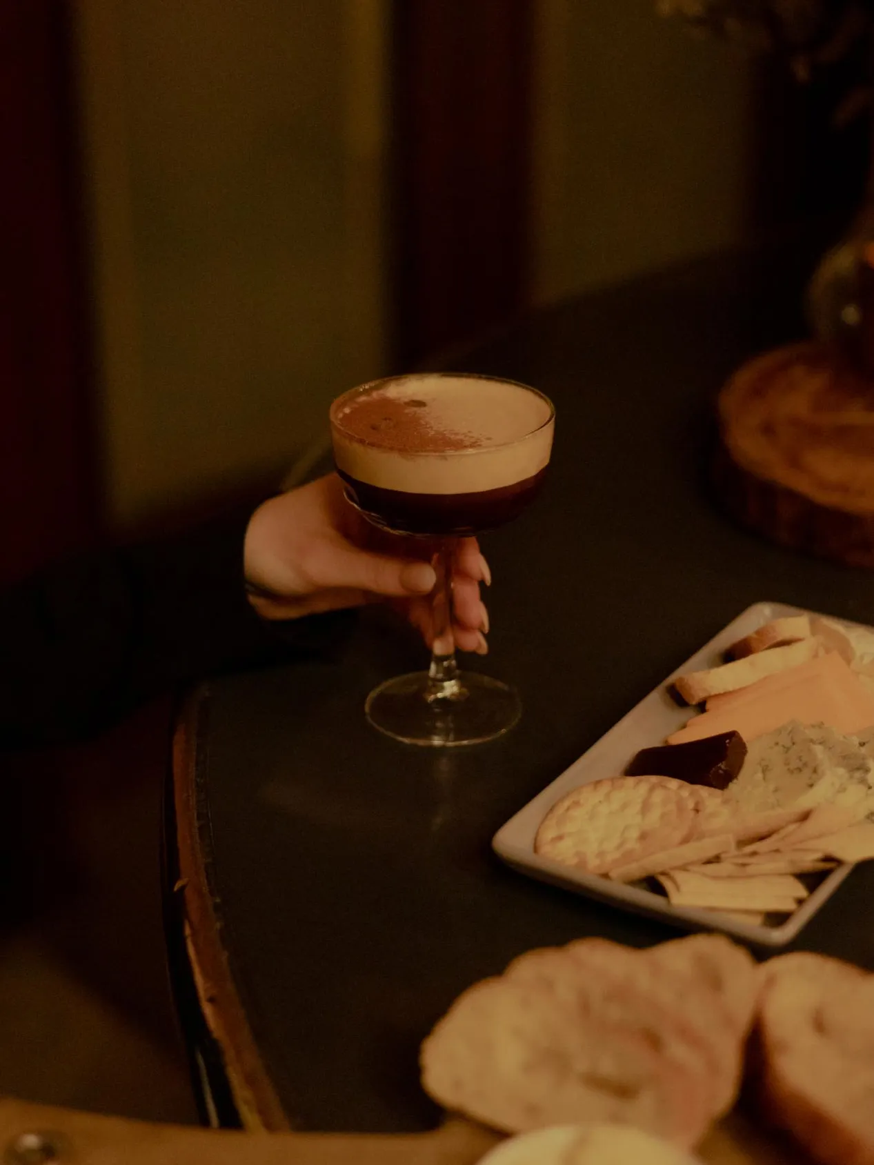 A hand holding an espresso martini on a table that also has a couple cheese plates.