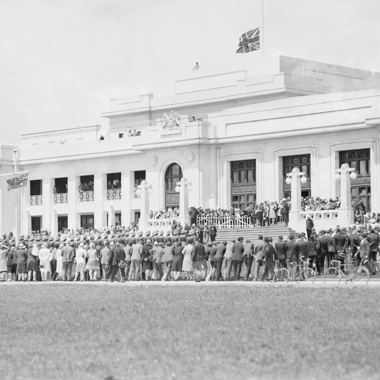 Black and white image of the front of Old Parliament House with a crowd of people of gathered at the foot of the stairs. The building is white with columns and an Australian flag flies at the top. 