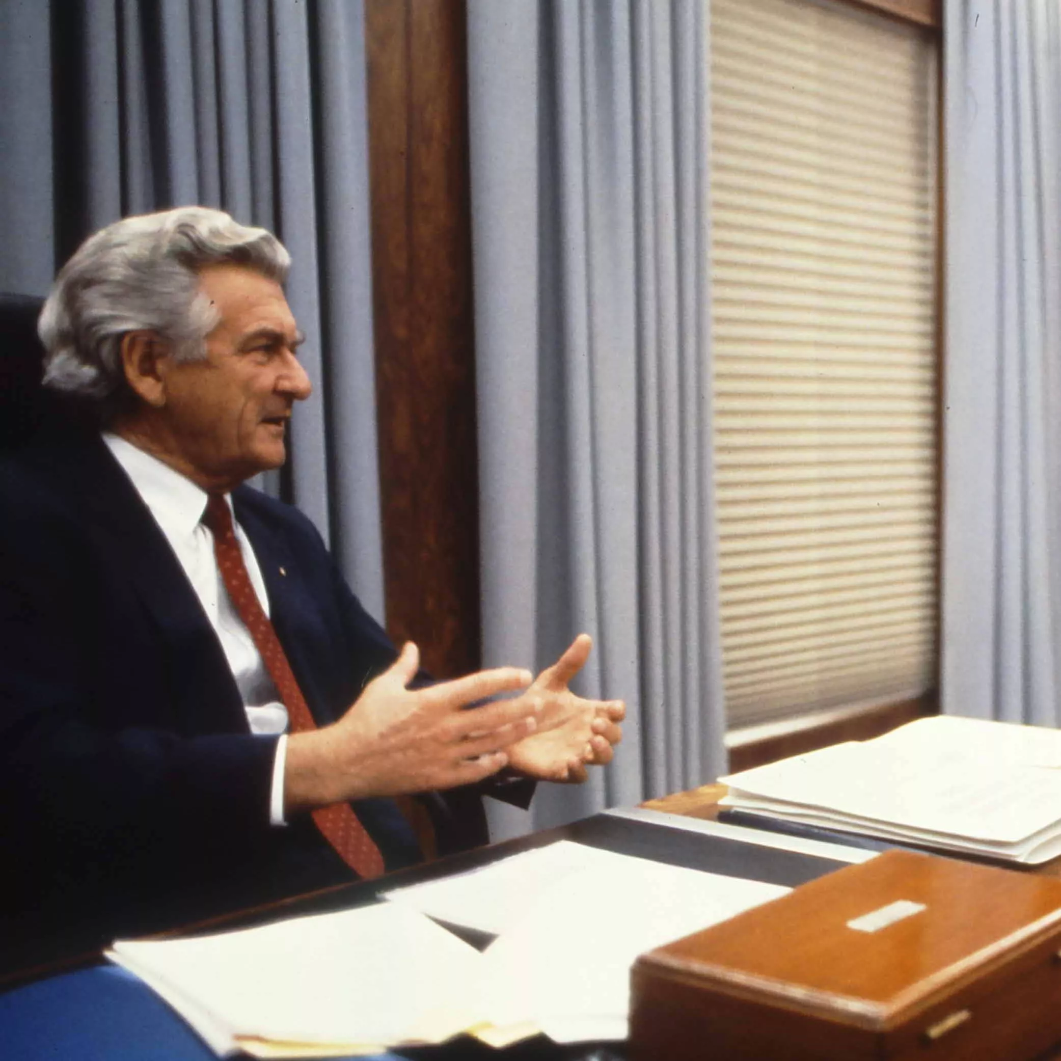 This colour photograph captures a conversation between Prime Minister Bob Hawke seated on the left and Treasurer Paul Keating seated on the right. The photo shows the corner of the desk with Hawke sitting behind the desk while Keating sits to the side with both men facing each other. Both are dressed in black suits with white shirts and red ties contrasting the pale blue curtains in the background.  