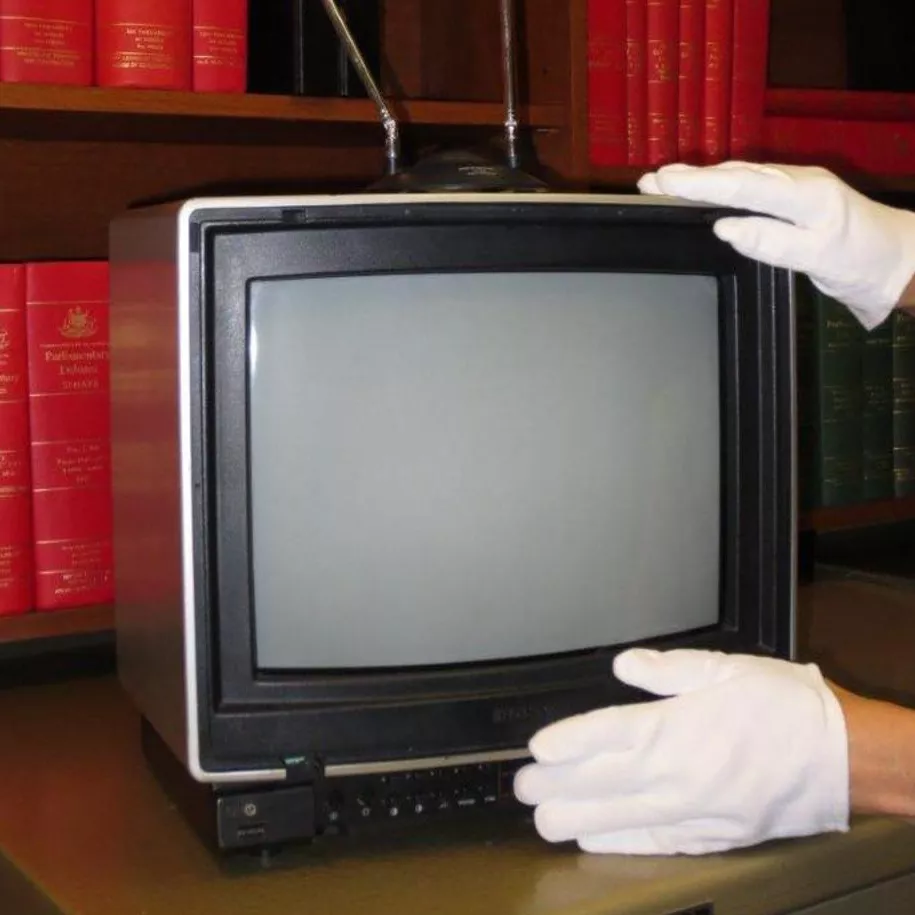 A pair of hands in white gloves positions a small television in front of a shelf with red books on it.