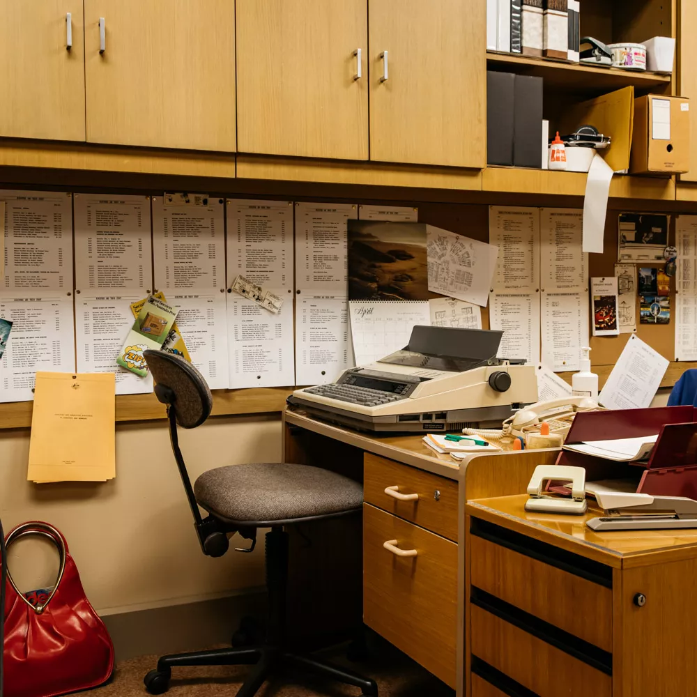 A 1980s looking office with brown desks, typewriters and printed pages stuck to a corkboard. 