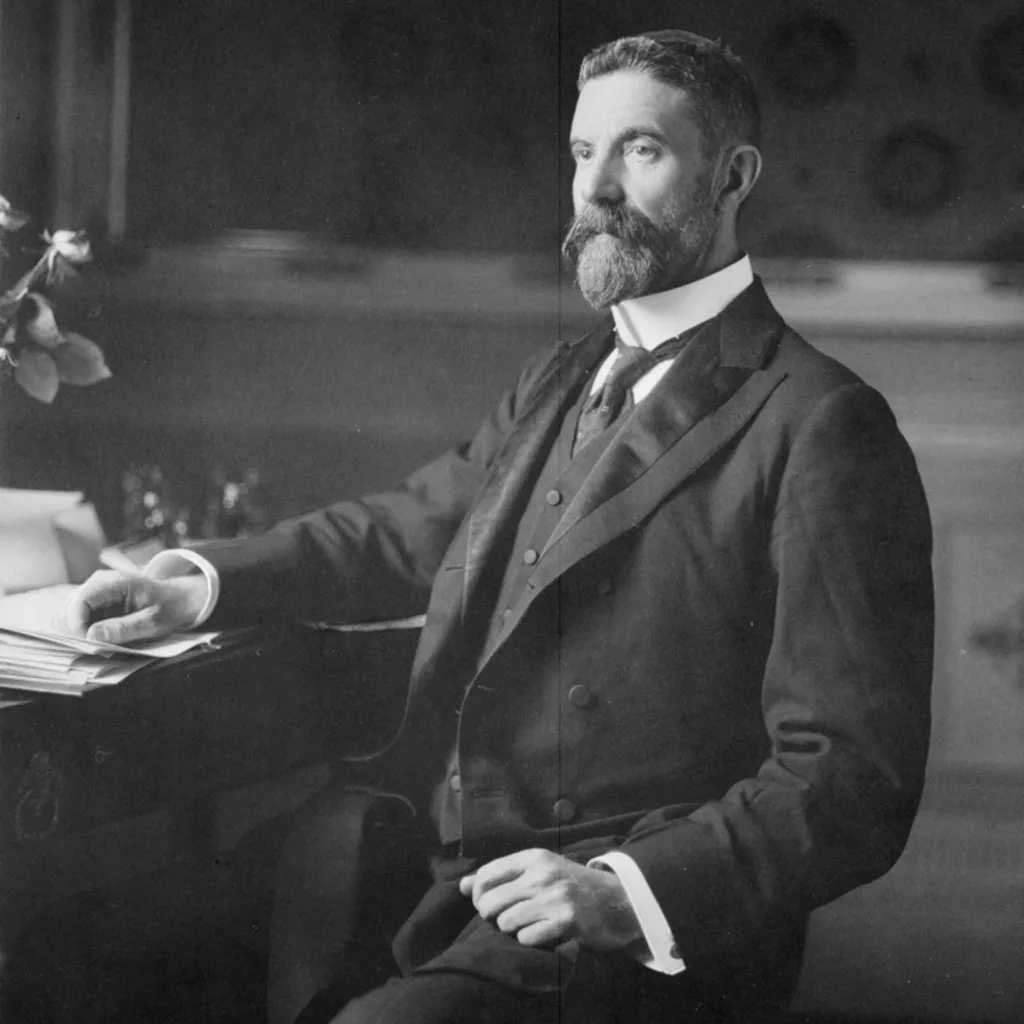 Alfred Deakin c.1901. Photograph National Library of Australia Alfred Deakin, wearing a three-piece suit and seated at a desk covered in paper documents.