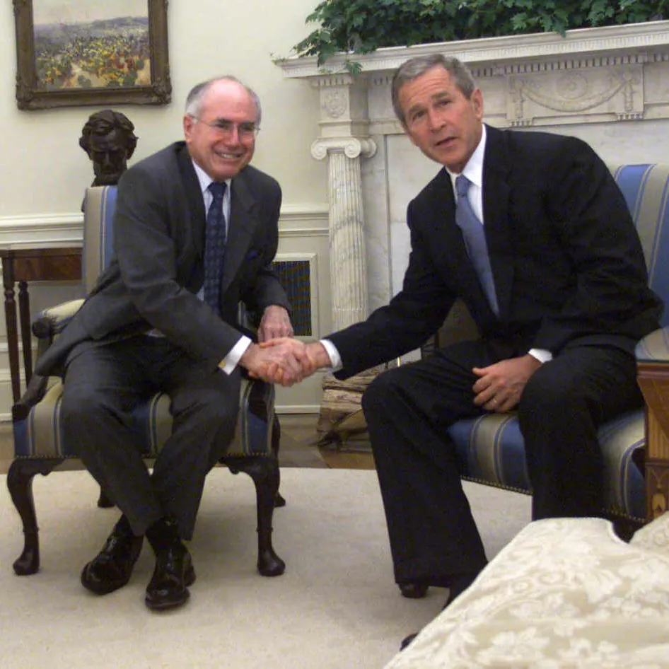 John Howard and George W Bush are seated in two plush chairs in front of a fireplace, smiling and shaking hands.