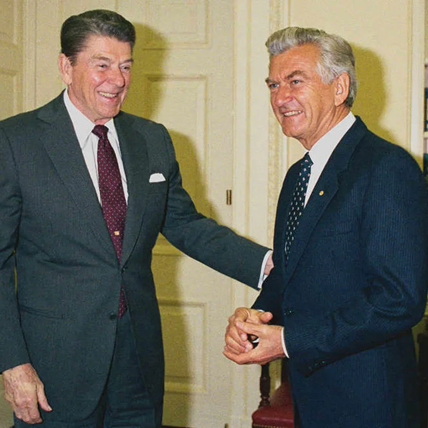 Ronald Reagan reaches an arm out to Bob Hawke, both smiling and wearing suits.