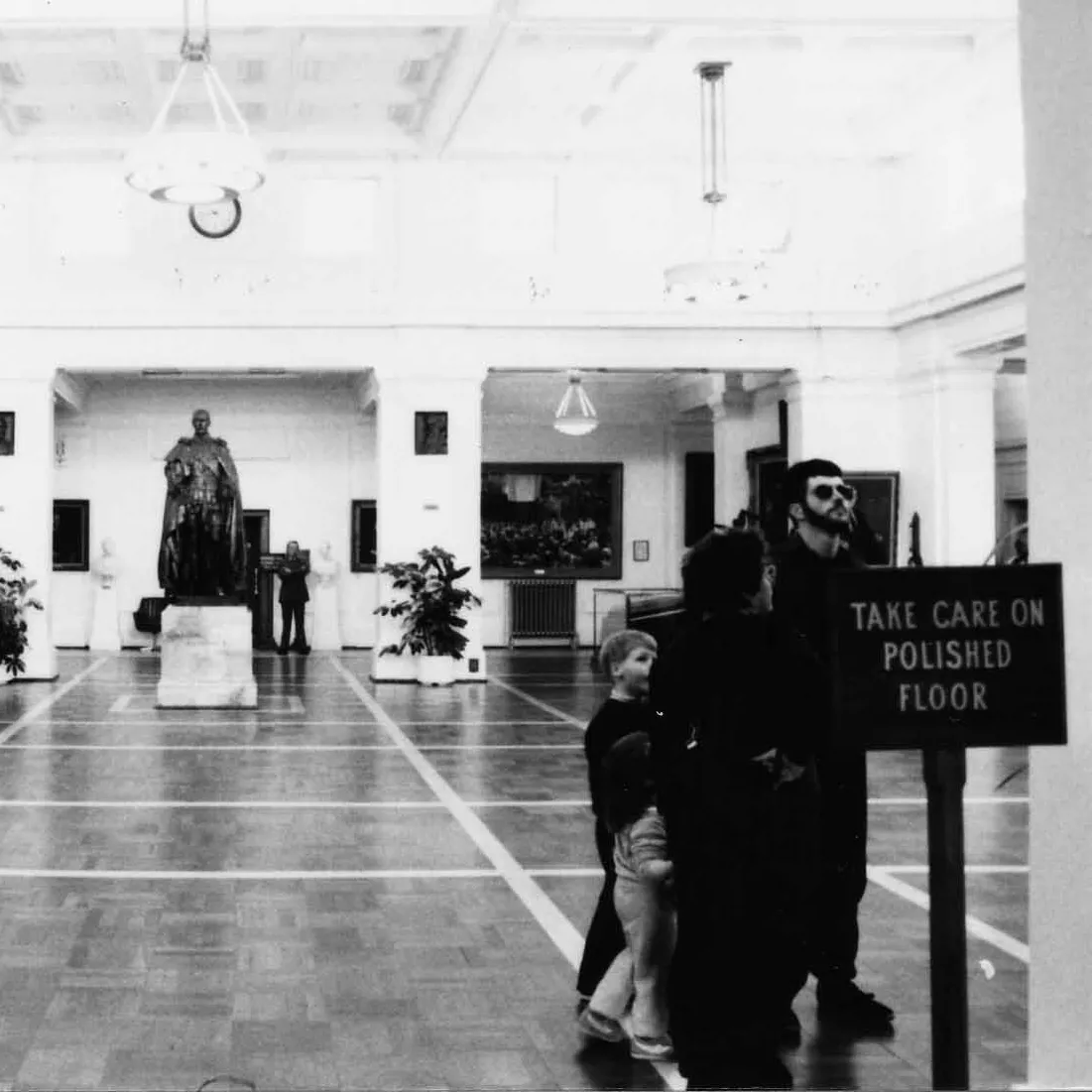 This black and white photograph of King’s Hall looks along the length of the room to the slightly larger than life bronze statue of King George the Fifth on a marble pedestal. King’s Hall is a large white room with a vaulted ceiling, celerestory windows, colonnades and a polished parquet floor.  The walls and columns feature artworks and portraits. You can see people wandering through the space including a family with young children and an older lady. In the foreground are two signs.