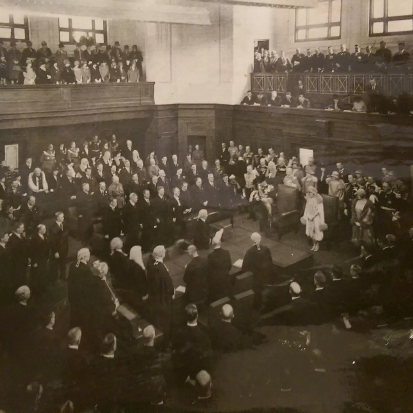 This black and white photograph shows the Duke of York reading a message from his father the King in the Senate Chamber. The room is crowded with at least 200 people, mostly men with some women all wearing their best. Men are dressed in suits, military uniform or gowns and wigs while the women wear formal dresses with hats and gloves. The crowd is in a horseshoe shape around the central table while the Duke stands at the head of the table.