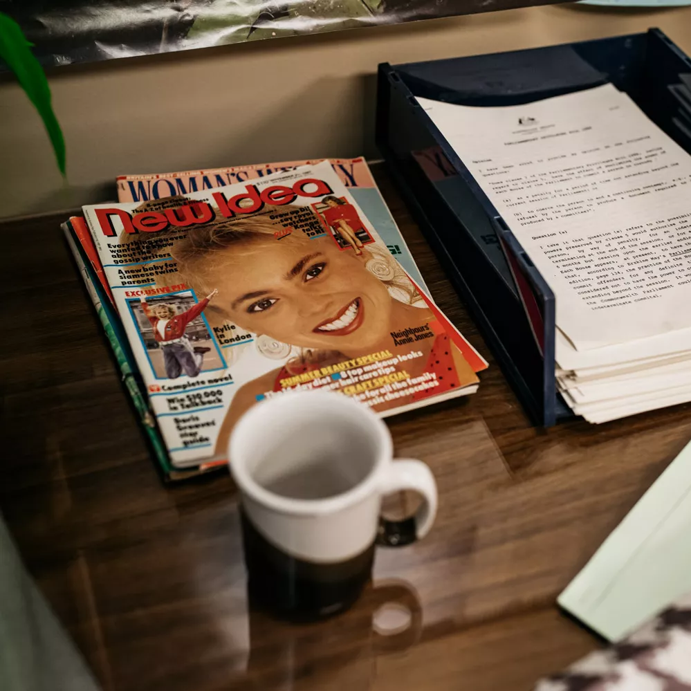 A staco of magazines and a coffee cup sat on one of the desks, alongside a tray of papers. On the wall behind, the bottom of a 'Kakadu National Park' poster can be seen.