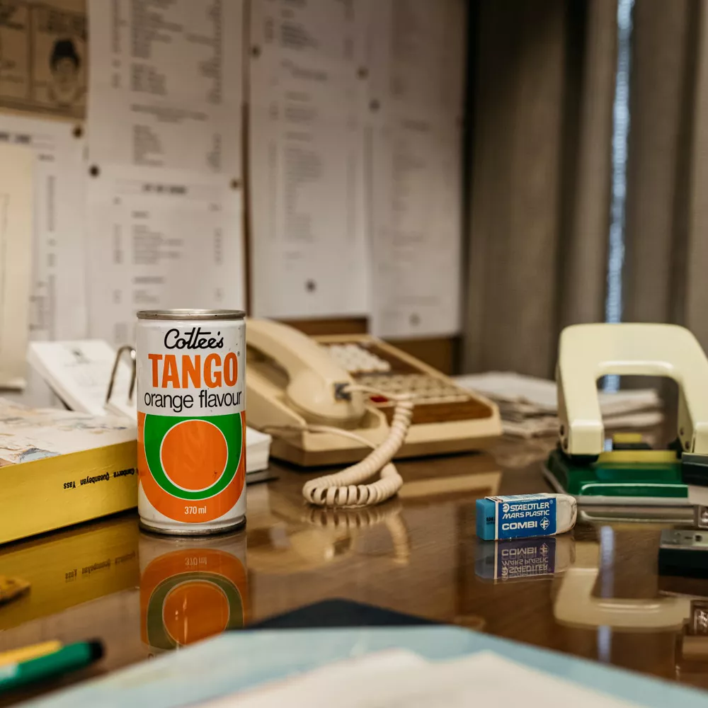 A can of 'Tango' drink sat on a desk, along with a telephone, hole punch and other typical clutter found on desks.