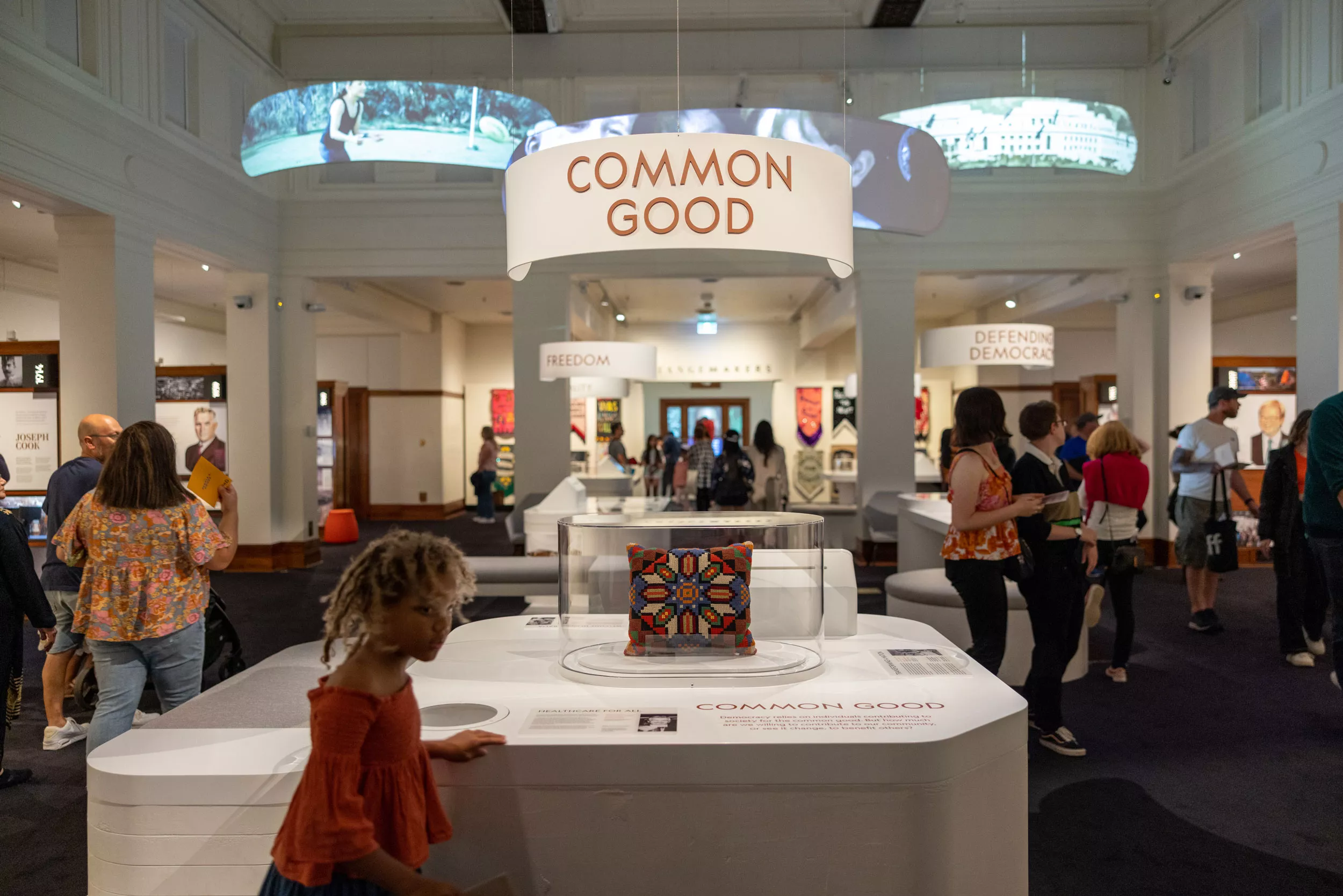 A wide shot of an exhibition with people walking around displays. Videos project on banners above and a sign that reads common good. 