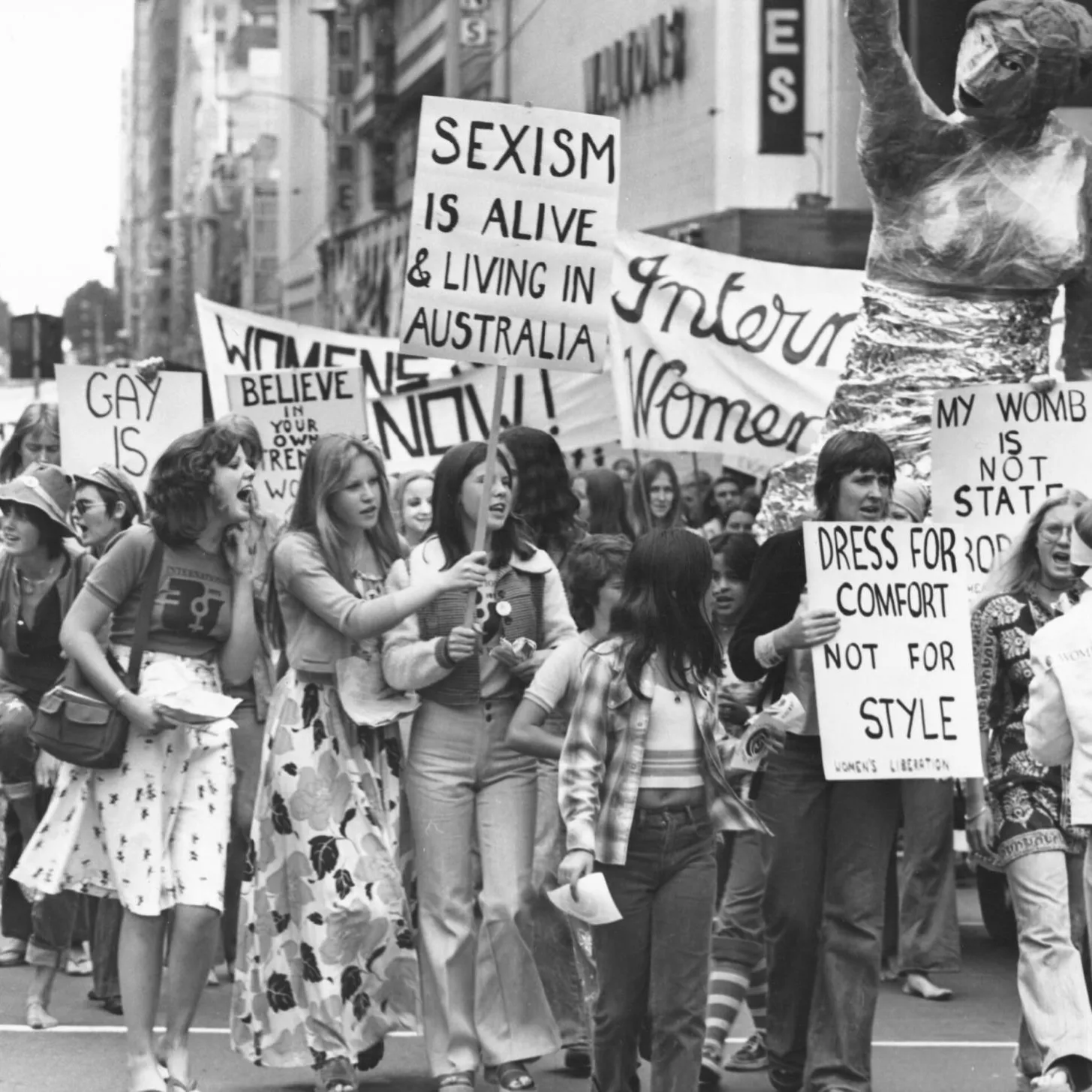 A black and white photo of a group of women holding protest signs in 1975. Signs say 'sexism is alive and well in Australia' and 'dress for comfort not style'.