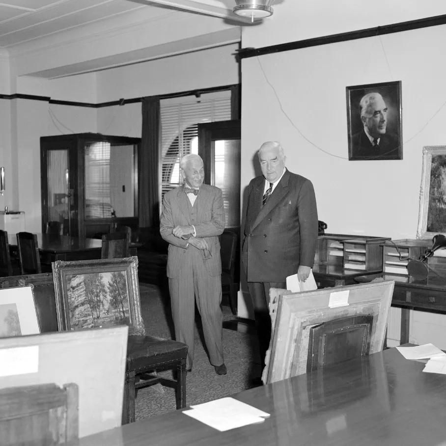 This black and white photograph shows two suited men standing side by side in the Government Party Room in 1962. On the left is Sydney businessman and philanthropist SH Ervin with a jaunty bow tie, on the right a more conservatively dressed Prime Minister Bob Menzies. They are surrounded by Australian paintings which are leaning against chairs, tables and walls. The two men are viewing the paintings that Ervin was donating to the National Collection. In the background are meeting tables and telephone booths