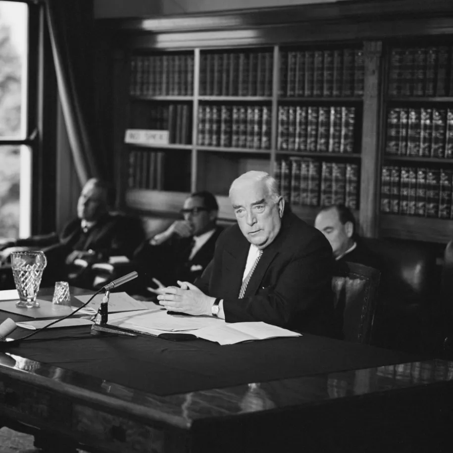 This black and white photograph shows Prime Minister Robert Menzies seated at a large table in the Government Party Room in 1965. He is conducting a press conference and there is a microphone, jug and water glass, and papers on the table. Behind him are three men seated in easy chairs and a bookcase filled with Hansards (volumes of parliamentary debates).  