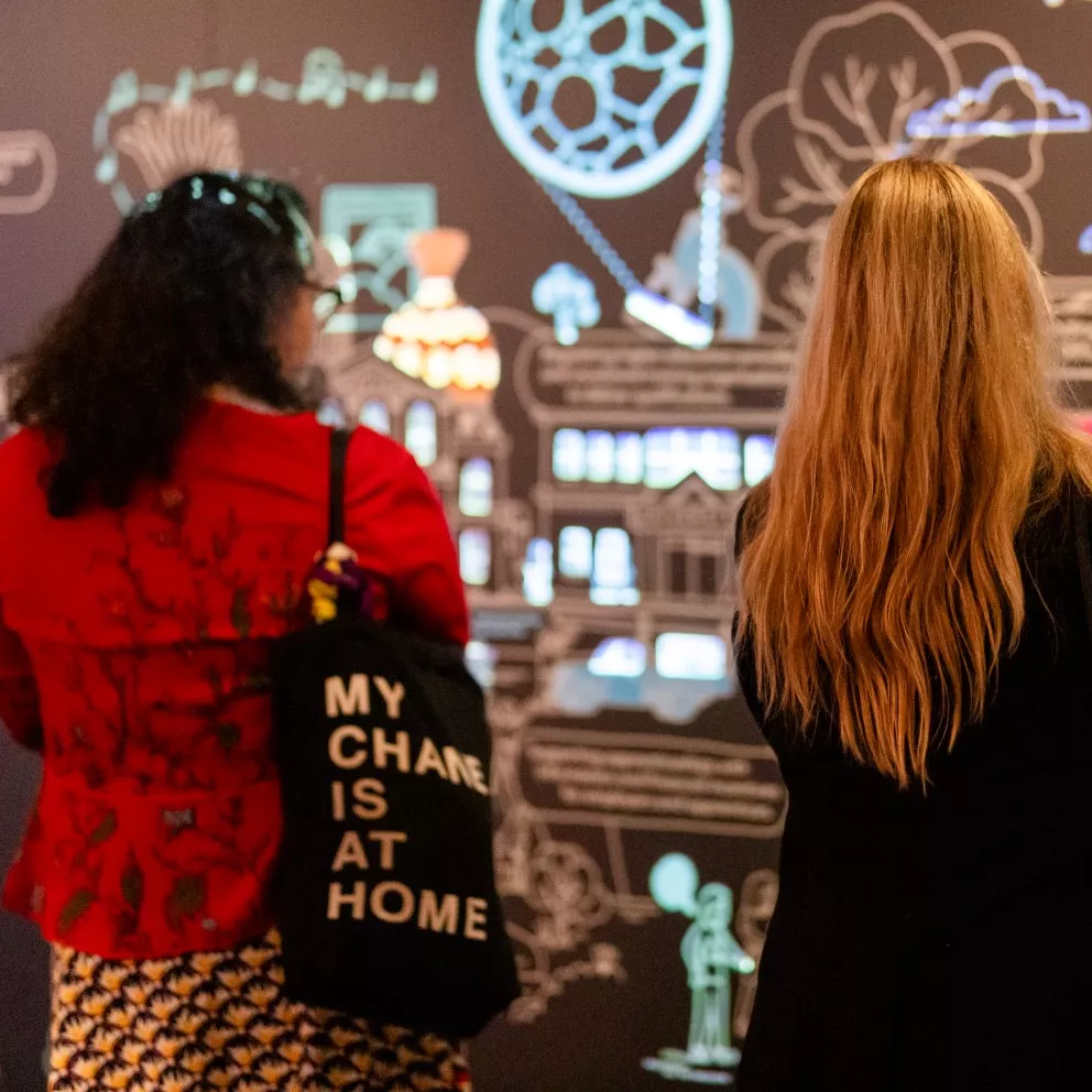 Two women look at a large wall installation of coloured lights and illustrations.