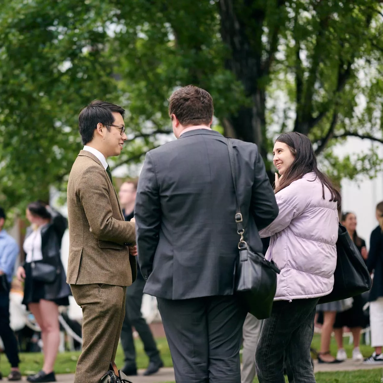 A group of three people talking together in the foreground, with many other people in the background, under a large tree in a courtyard.