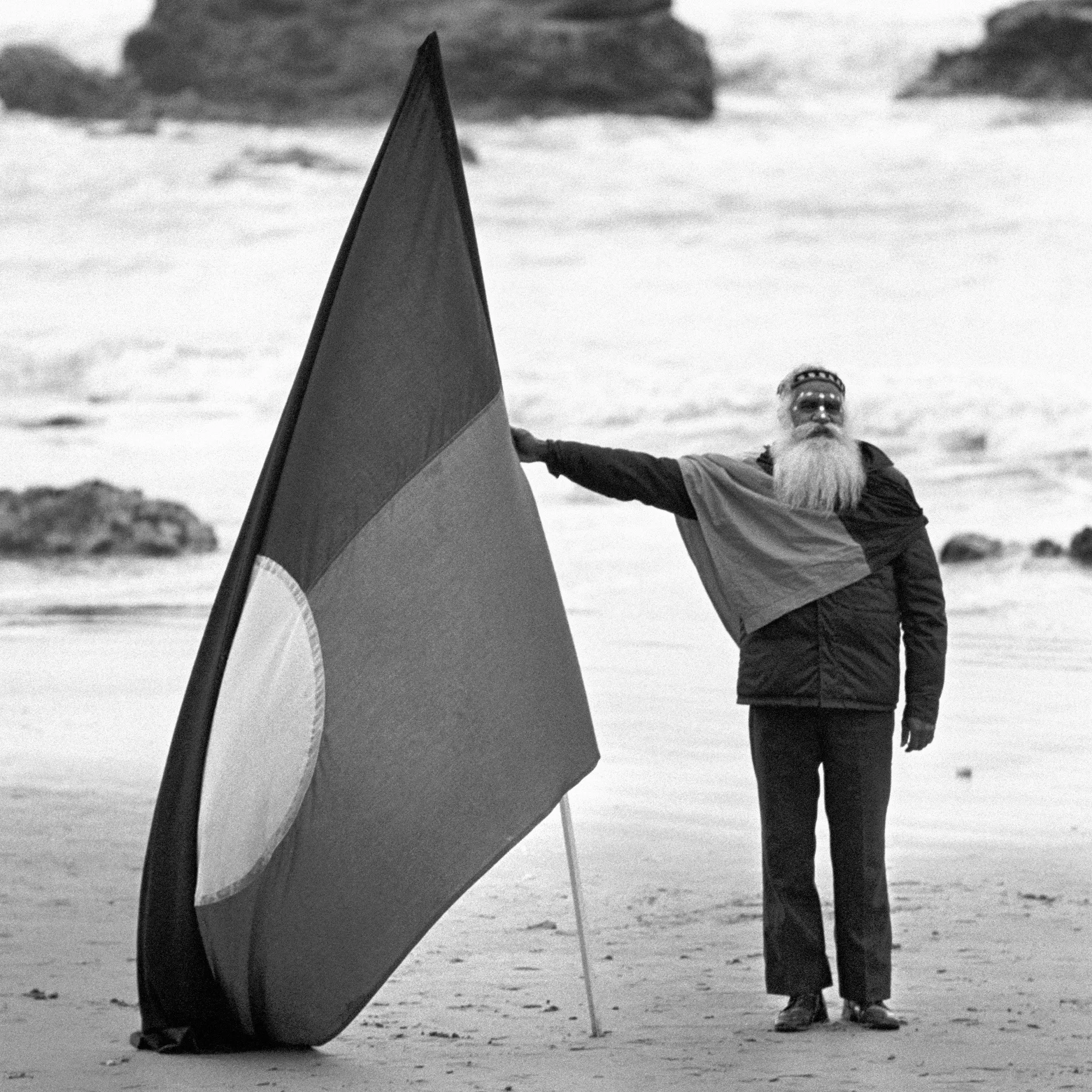 Burnum Burnum, a man with a large white beard, stands on the beach holding a large Aboriginal Flag.