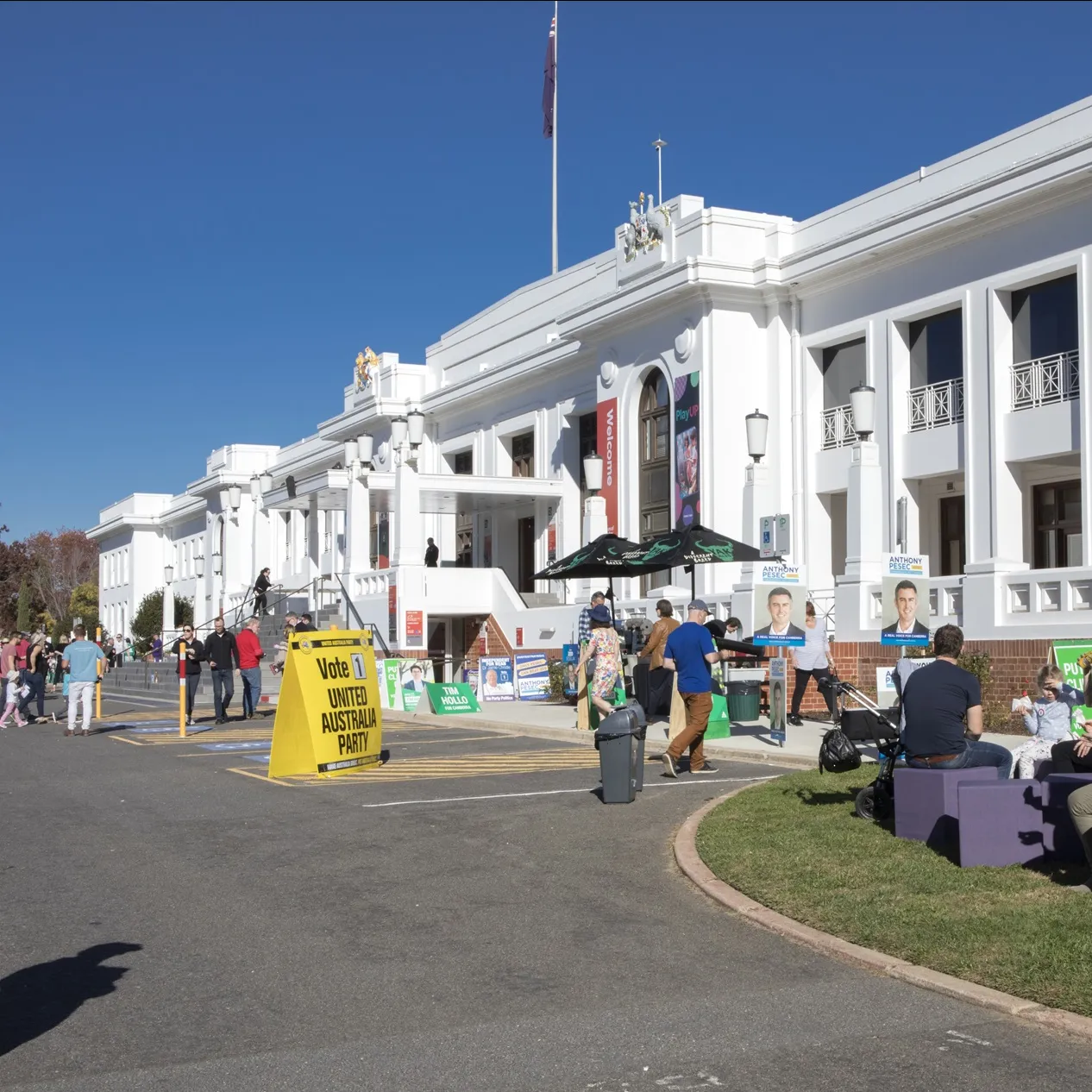 Many colourful signs and placards displaying political candidates, as people enter and leave Old Parliament House, with some sitting on purple blocks on the front lawn.