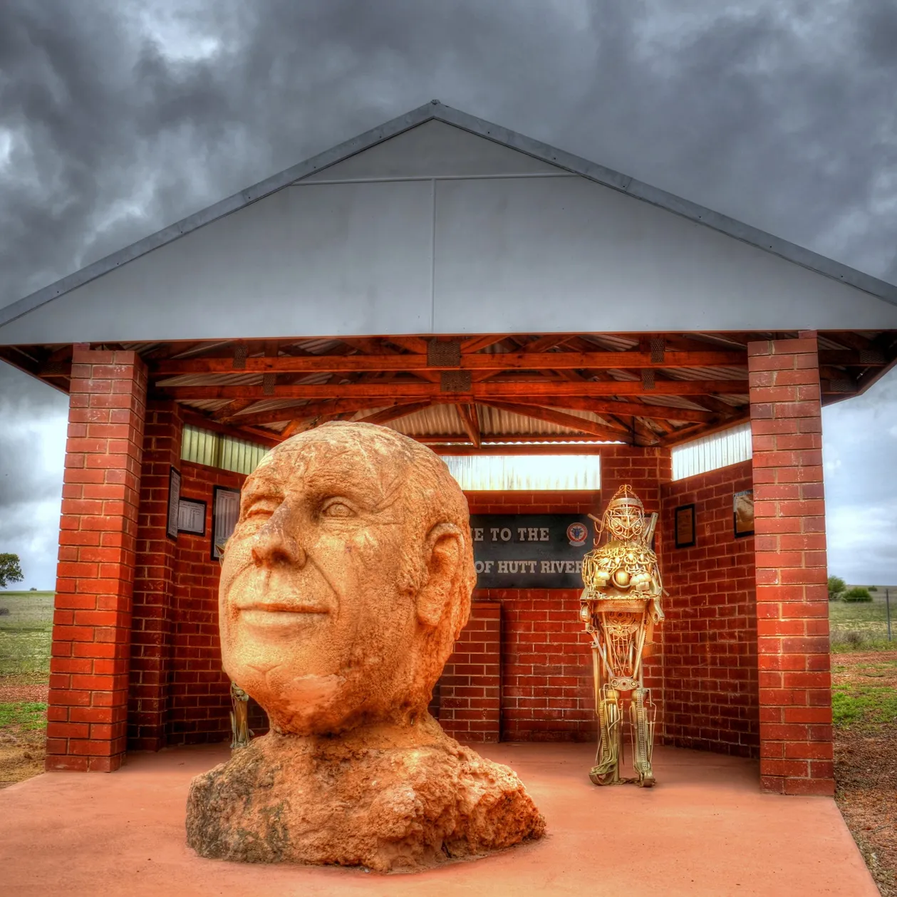 A hand-carved rock bust depicting the head and shoulders of 'Prince' Leonard George Casley, positioned in front of a redbrick shelter in a paddock.