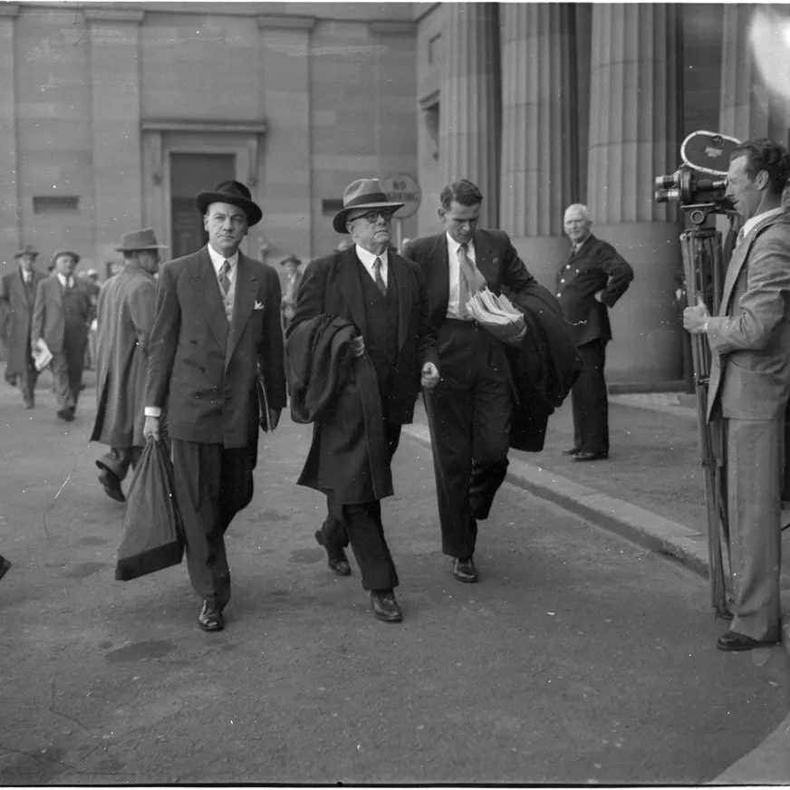 Dr Evatt is flanked by two assistants as he enters the Petrov Royal Commission in Darlinghurst. Men are walking nearby, with a man standing with a camera recording them.