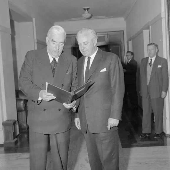 Prime Minister Menzies is holding a document, which both he and Harold Holt are looking at.