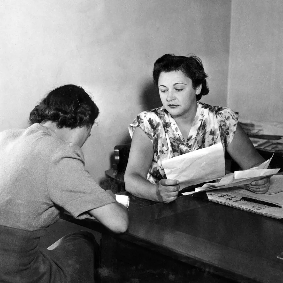 Nancy Wake is at a desk holding papers. She is looking across at another woman who is writing.