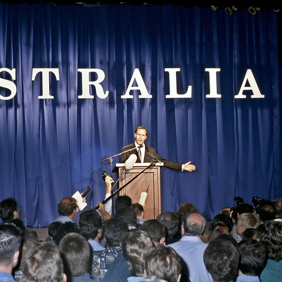 Paul Keating stands at a lecturn with the word 'Australia' behind him. A crows 