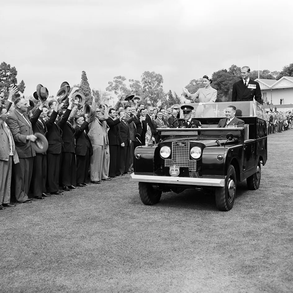 The Queen and Prince Phillip are standing in the back of a car waving to a crowd who are all waving their hats to them.