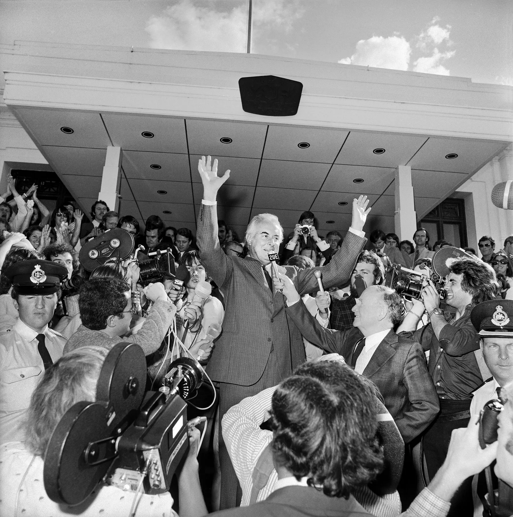 Gough Whitlam stands with his hands in the air amidst a crowd of reporters with microphones and cameras on the steps of Old Parliament House on November 11 1975.