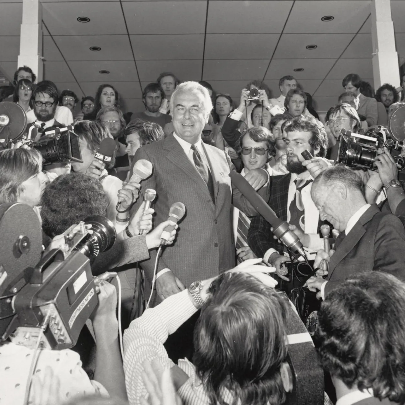 Gough Whitlam on the steps of Old Parliament House surrounded by the press who are holding microphones and video cameras on 11 November 1975.