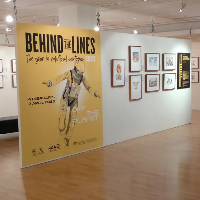 A gallery space with framed cartoons on the walls. A large, yellow introductory panel features the illustration of an astronaut and the words 'Behind the Lines'.