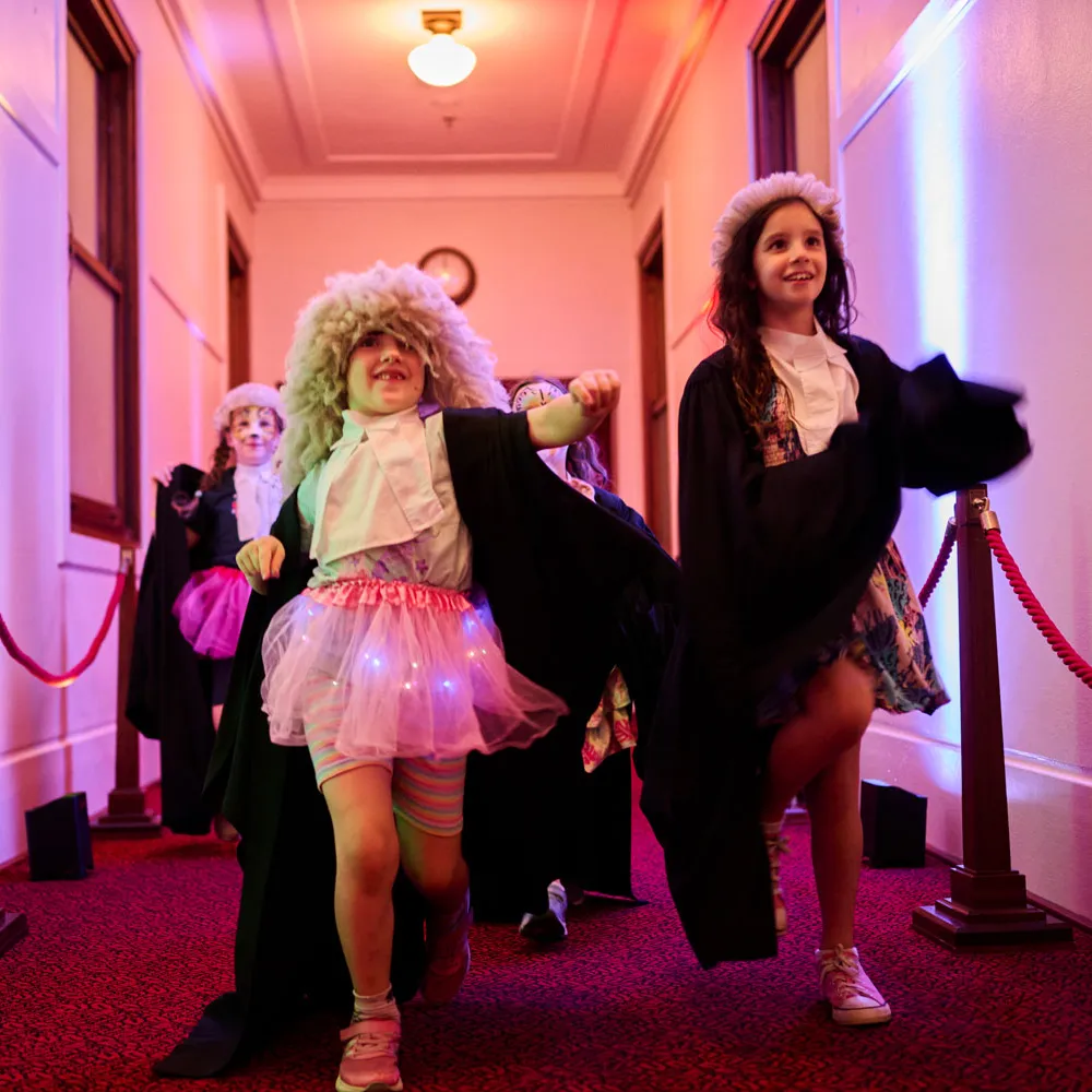 Two girls dressed up in costume white wigs and black gowns walk down a hallway with red carpet in Old Parliament House. One of the girls is also wearing a pink tutu with LED lights in it.