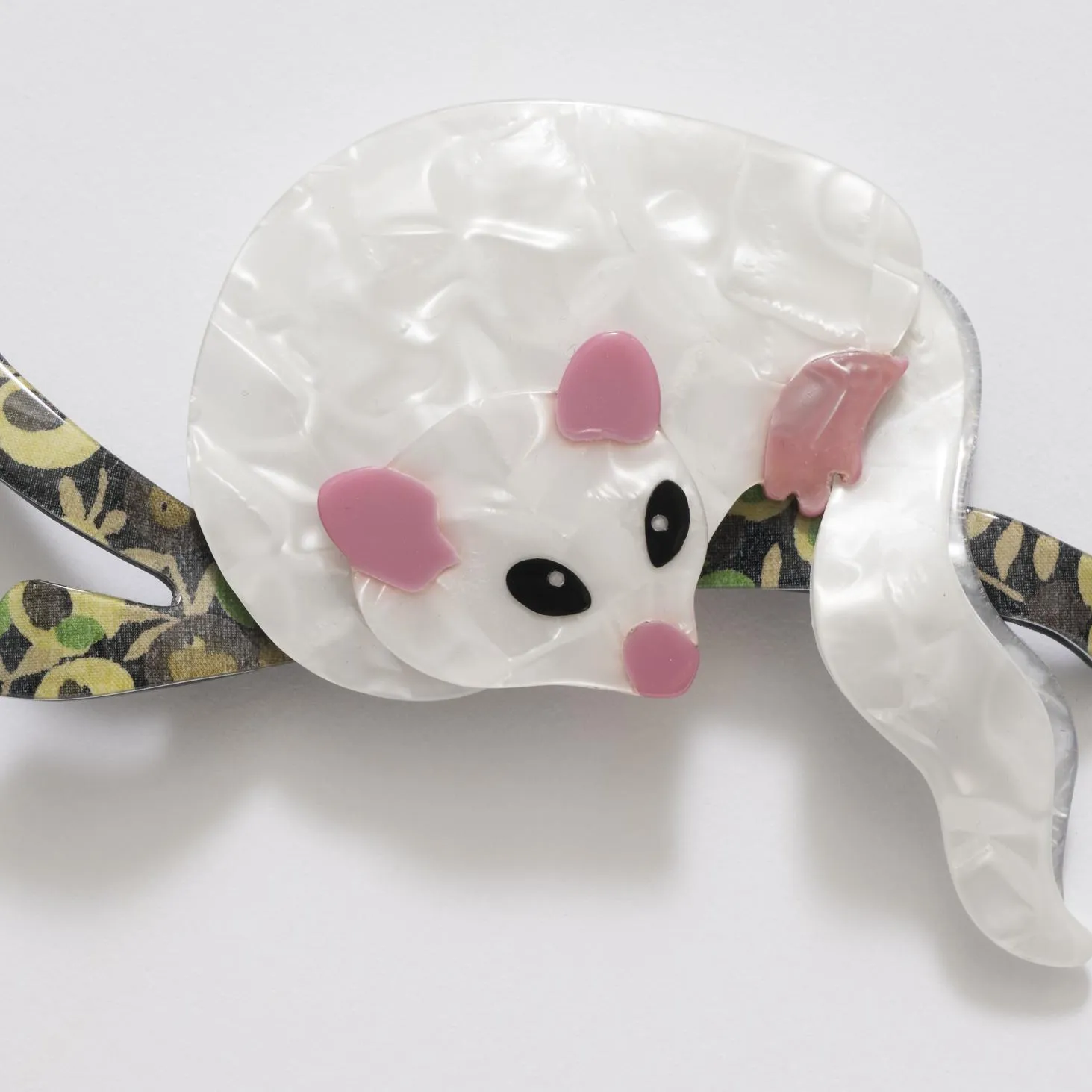 A celluloid possum brooch. The possum is white with pink ears, nose and feet, it sits on a black, green and gold branch.