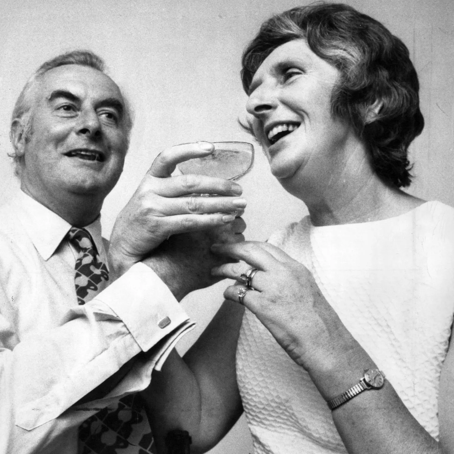 Gough Whitlam and Margaret Whitlam smile and hold a coupe of champagne between them.