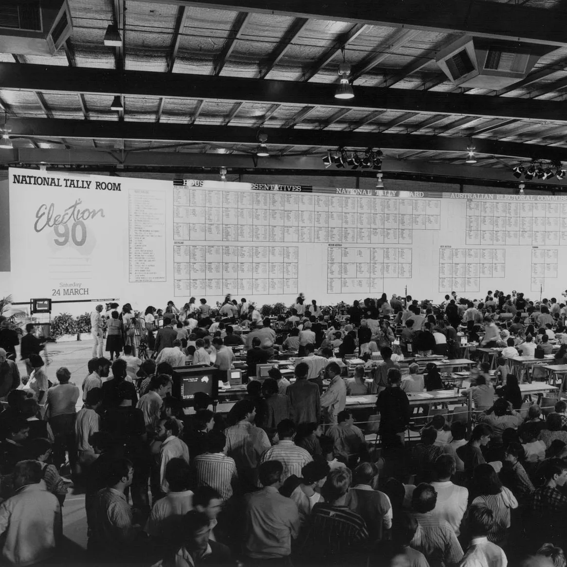 A crowd of people stand in the National Tally Room in Canberra looking at the giant National Tally Room board showing the results of the 1990 election.