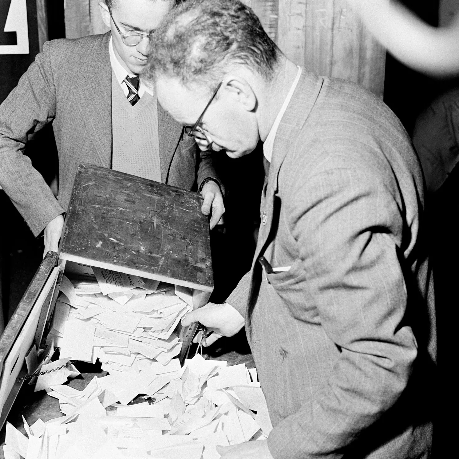 Two men empty folded votes out of a wooden ballot box for the 1954 election.