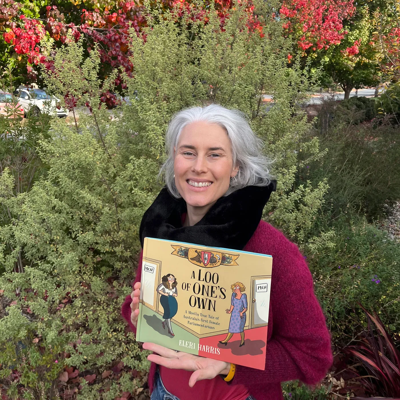 A woman holds a book titled A Loo of One's Own in front of autumn foliage. A woman holds a book titled A Loo of One's Own in front of autumn foliage.