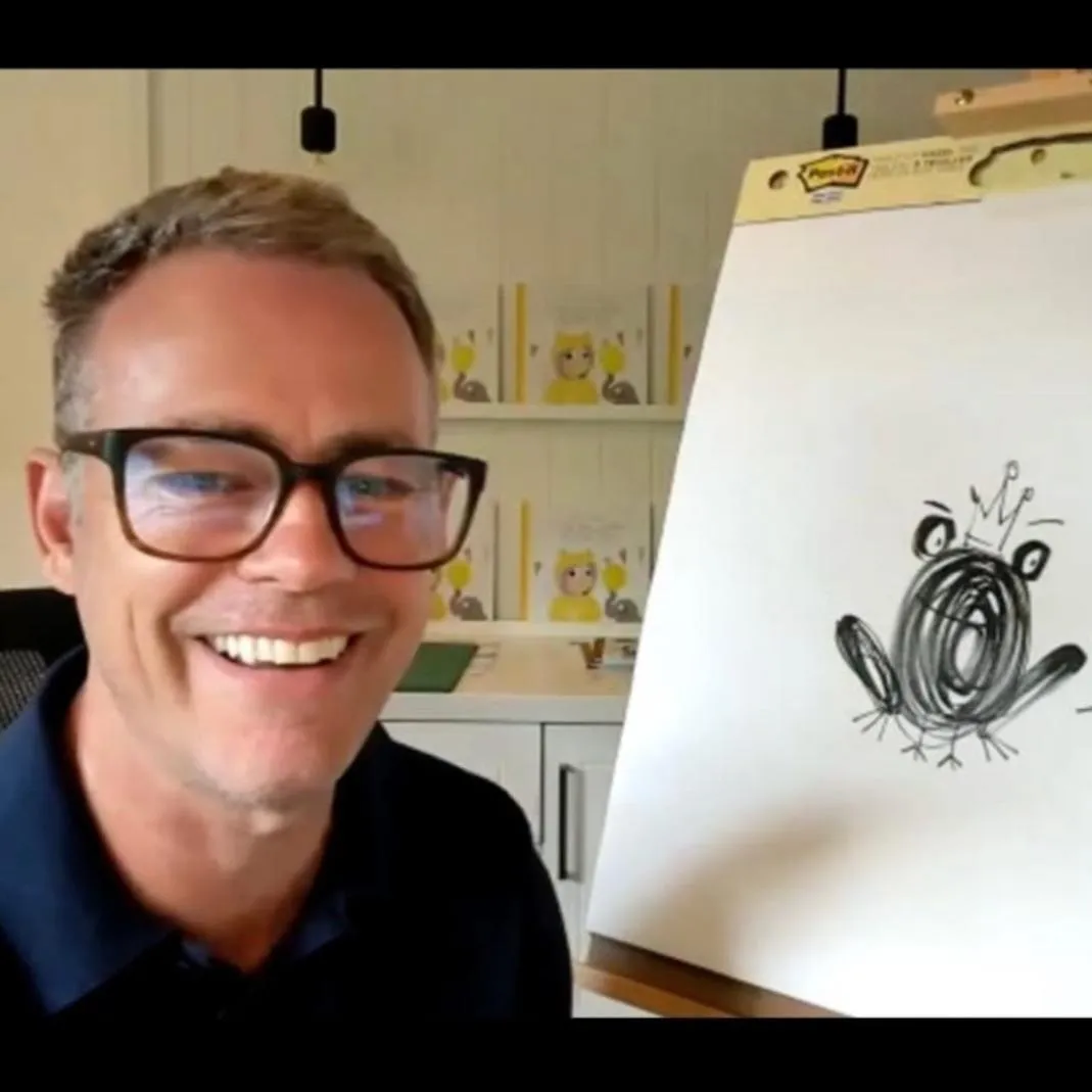 A man wearing glasses is smiling at the camera. There is a drawing next to him on an easel.