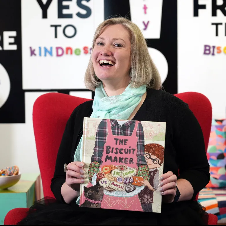 A woman sitting holding a picture book titled 'The Biscuit Maker' by Sue Lawson, illustrated by Liz Anelli