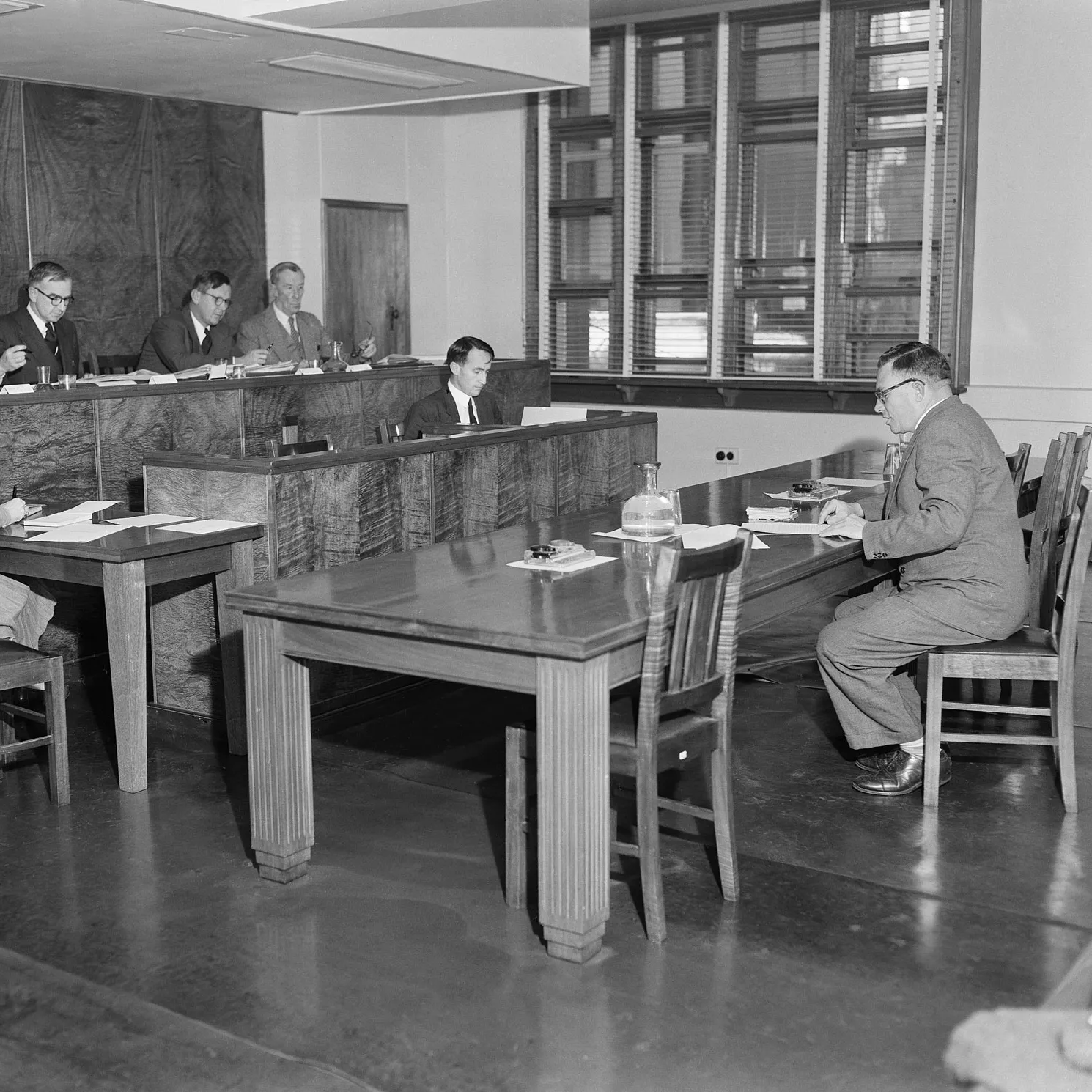 A man reading papers of evidence to a room of a couple people that make up the Royal Commission