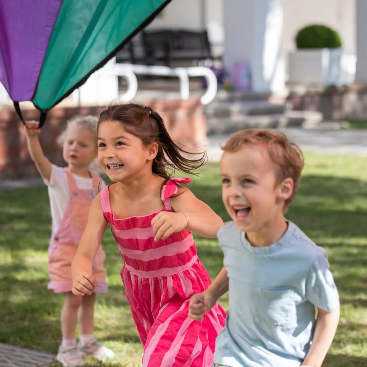 Three kids in bright colours run underneath a multi-coloured parachute in the gardens of Old Parliament House.
