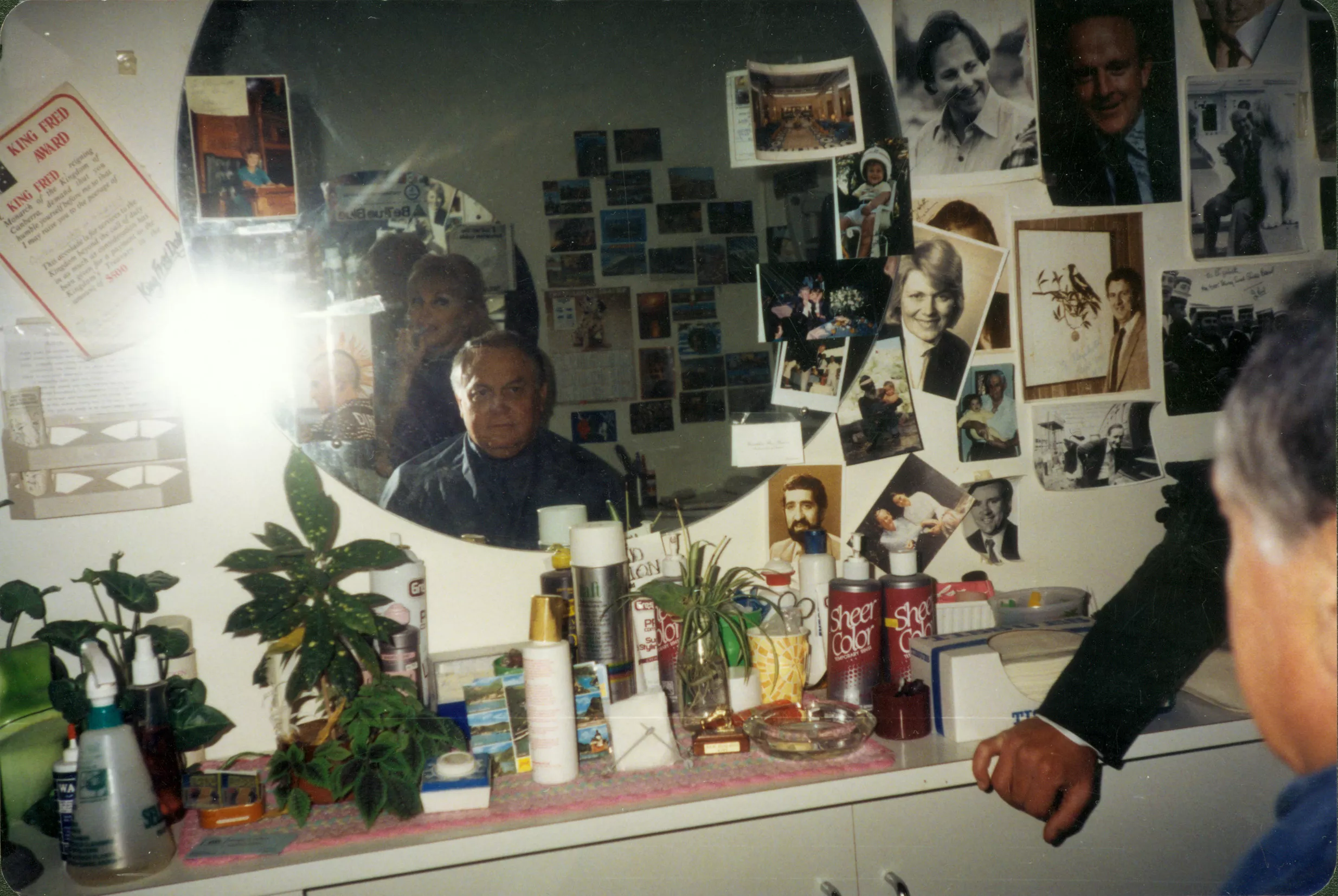 This colour photograph depicts the inside of Lizzie Scott’s salon. Gary Nehl MP sits in the barber’s chair with Lizzie standing directly behind him. They  are reflected in a circular mirror which is mounted on the wall. Under the mirror, there is a counter which is littered with pot plants, hair spray, tissues, scissors, spray bottles, creams, postcards and an ashtray. Surrounding the mirror, are photographs of people in colour and black and white. 