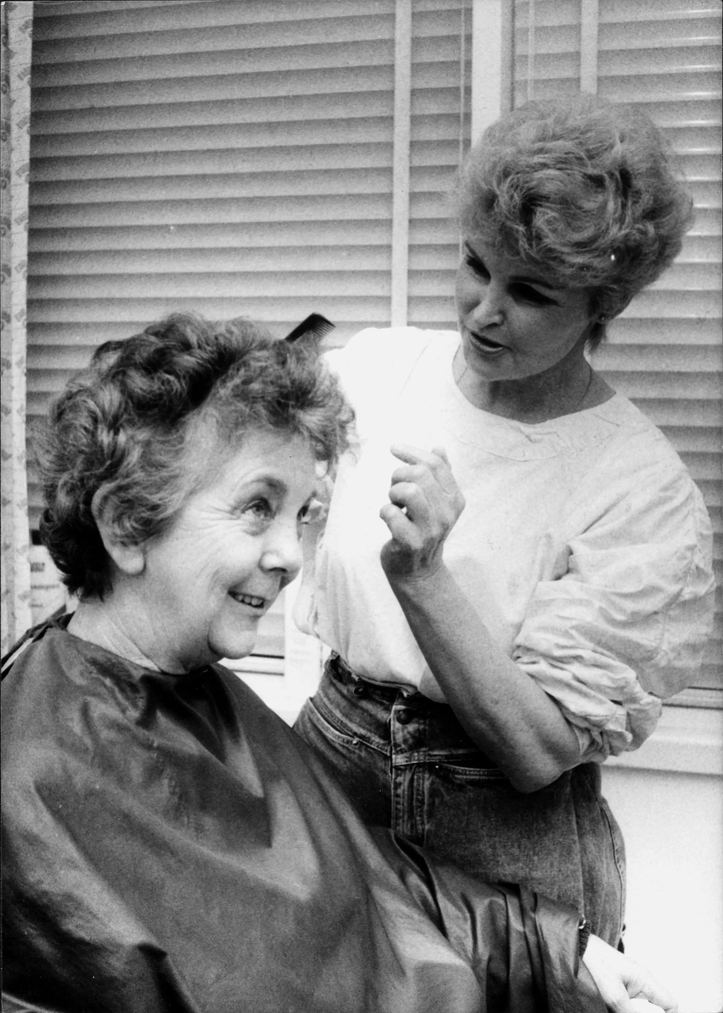 This black and white photograph depicts hairdresser Lizzie Scott, arranging Speaker Joan Child’s hair. Joan is sitting in the barber’s chair and is covered with a dark  cape while Lizzie stands to her left wearing a white shirt and jeans and holding a black comb.  