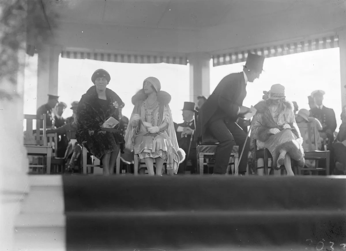 This black and white photograph lets us peek over the edge of the carpeted floor of the white timber Royal dais constructed at York Park for the official party to watch the Troop Review. On the left, a seated Mrs Ethel Bruce chats with the Duchess of York. Ethel is dressed in a dark, luxurious fur coat with cloche hat and gloves. The Duchess is richly dressed in a pale-coloured lustrous dress with elaborate embroidery and a fur-trimmed cape, cloche hat and white gloves. Prime Minister Bruce, in top hat and 