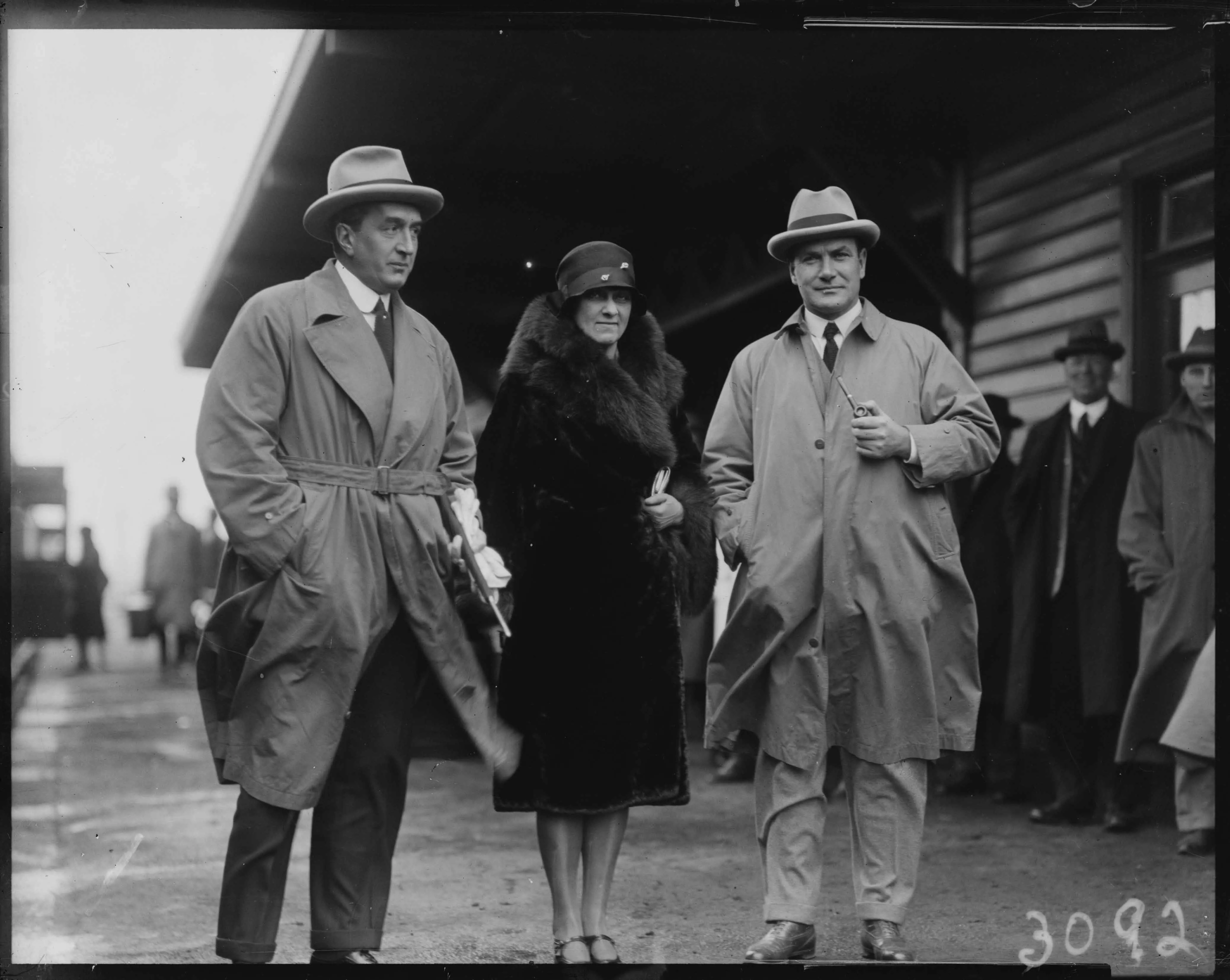 This black and white photograph shows three individuals standing on the platform at the weatherboard Canberra Railway Station on 8 May 1927. In the distance you can see the open doors of a train and people walking down the platform with luggage. Some other people are sheltering close to the railway station building. The weather looks cool and windy. On the left is Prime Minister Stanley Melbourne Bruce dressed in a dark suit with white shirt and dark tie over which is a belted trench coat in a lighter shade