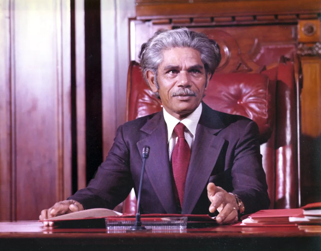 Neville Bonner seated in the Senate chamber, wearing a suit and red tie.