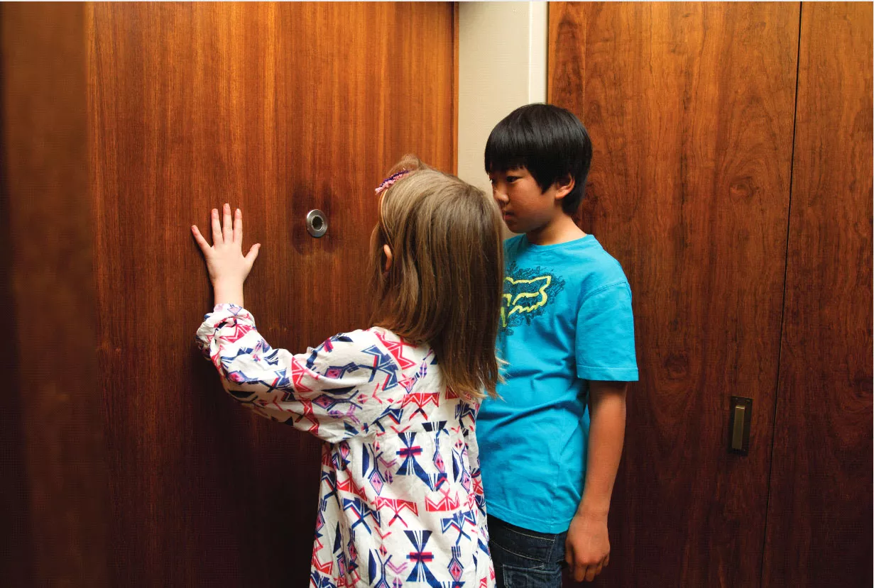 Two children investigating what appears to be a spy-hole in a wooden wall