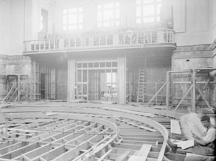 This black and white photograph depicts the House of Representatives Chamber partway through construction. The horseshoe shape of the chamber seating is visible in the timber framework. The public gallery is partly constructed and westerly light pours through the high windows. There are timber frames and a ladder against the back wall of the chamber. In the right foreground, a lone construction worker sits between the timber floor beams, reading a newspaper and smoking a pipe. 